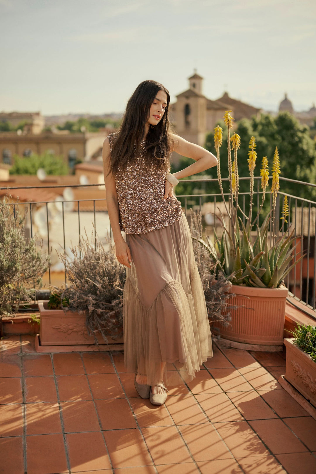 Femme portant un haut à paillettes et une jupe fluide, posant dans un décor ensoleillé sur une terrasse.