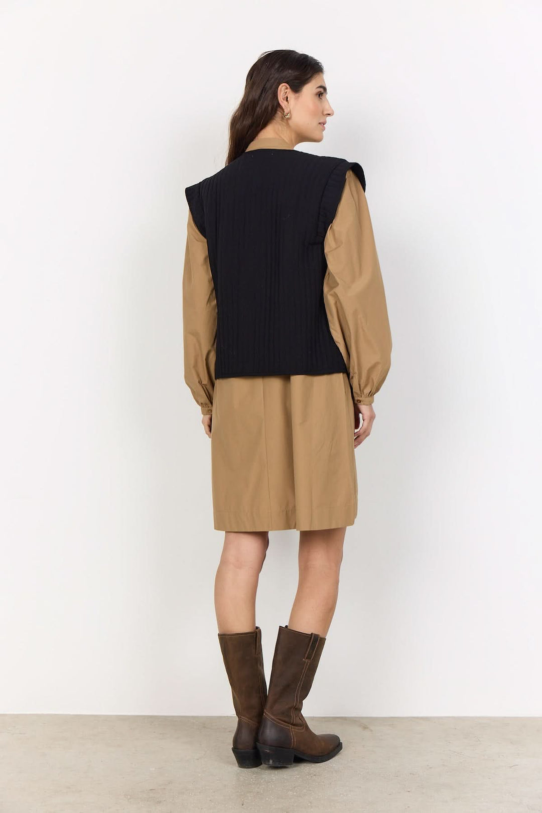 Veste courte sans manches, encolure ronde, portée avec une robe beige et des bottes marron pour un look décontracté.