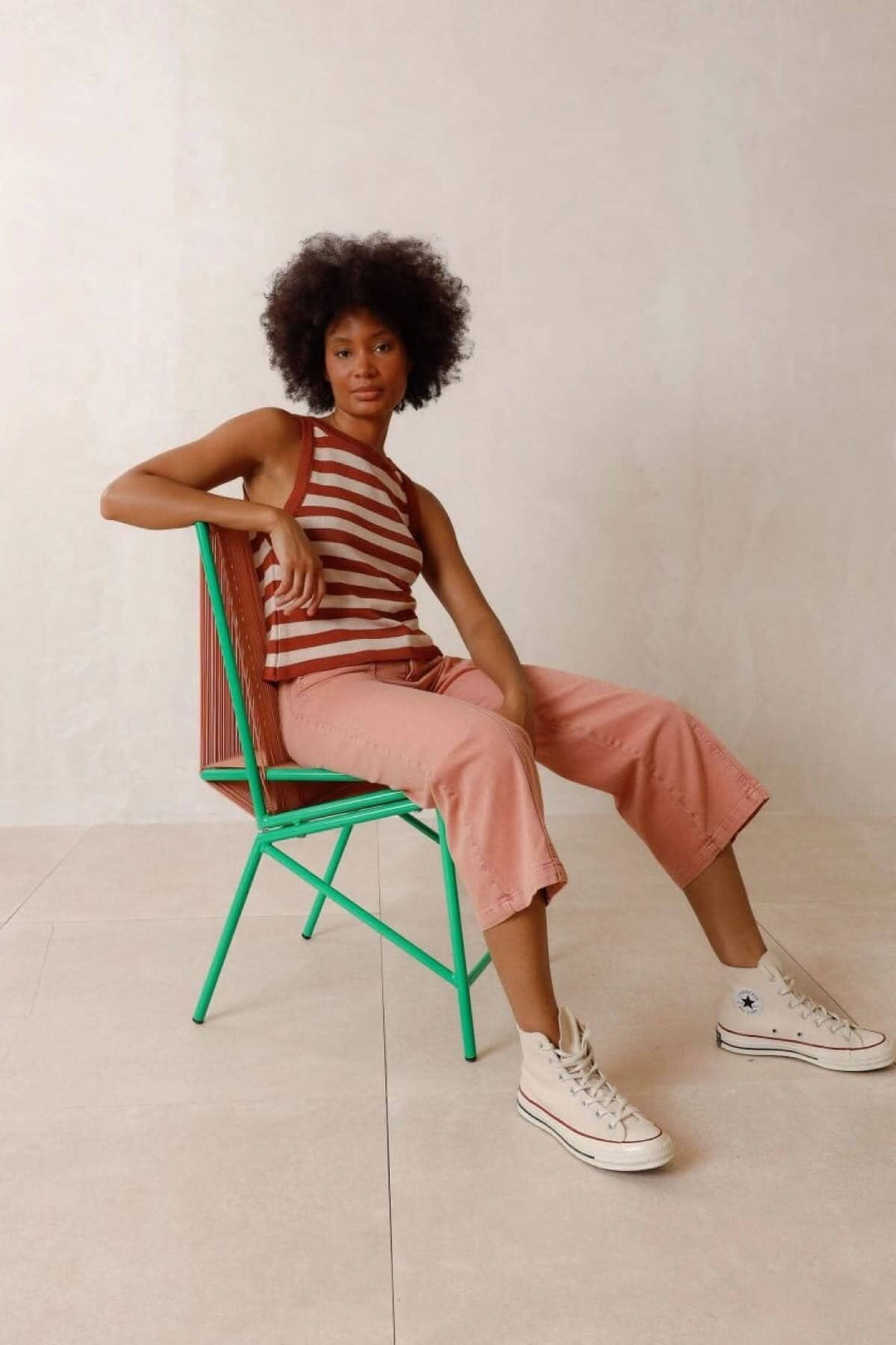 Femme portant un haut rayé à bretelles et un pantalon rose, assise sur une chaise verte.