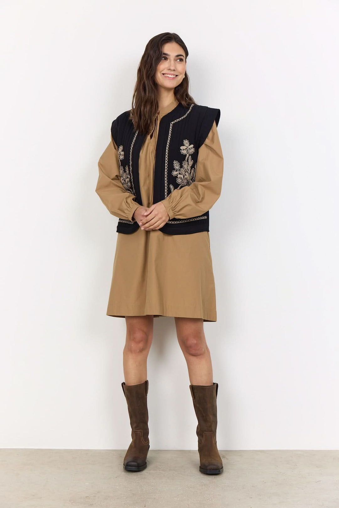 Veste courte sans manches noire avec broderies florales, portée avec une robe beige et des bottes marron.