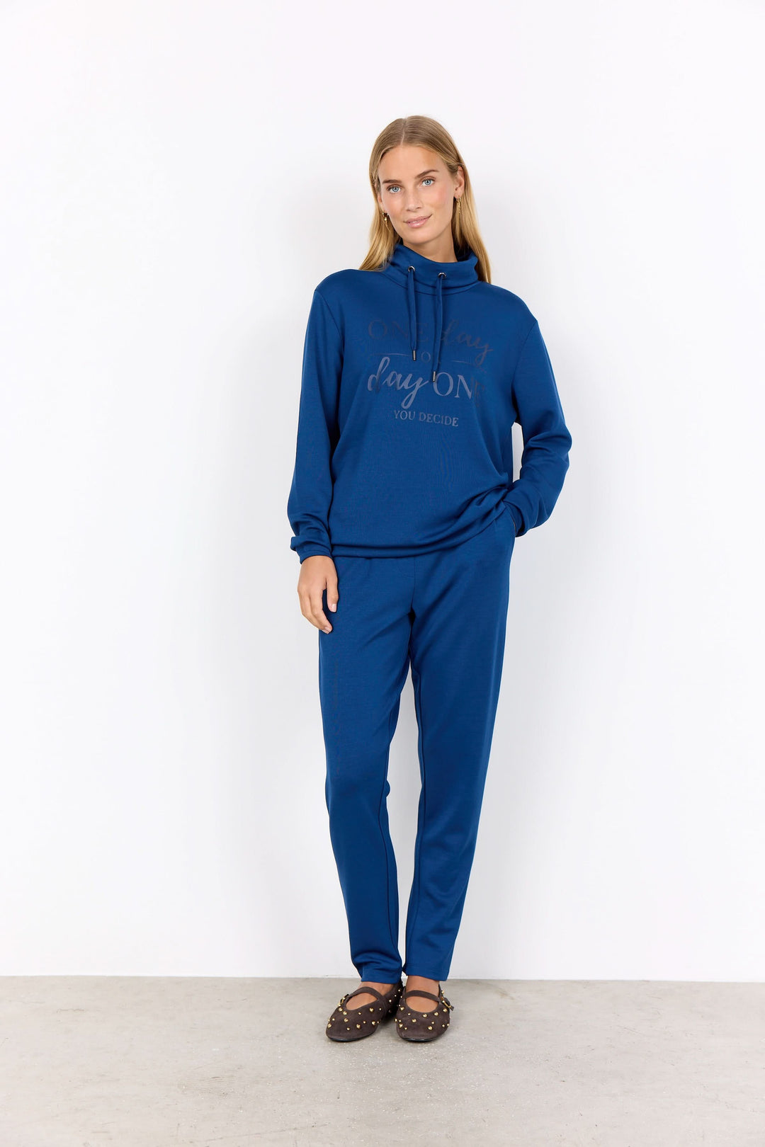 Chandail col montant bleu avec message positif, porté avec un pantalon assorti pour un look décontracté.