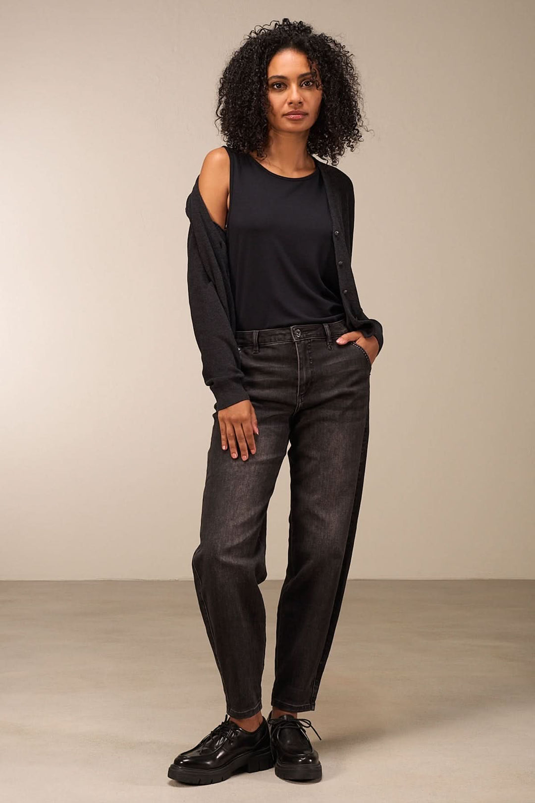 Femme portant un cardigan simple, t-shirt noir et jeans gris, dans un décor minimaliste.