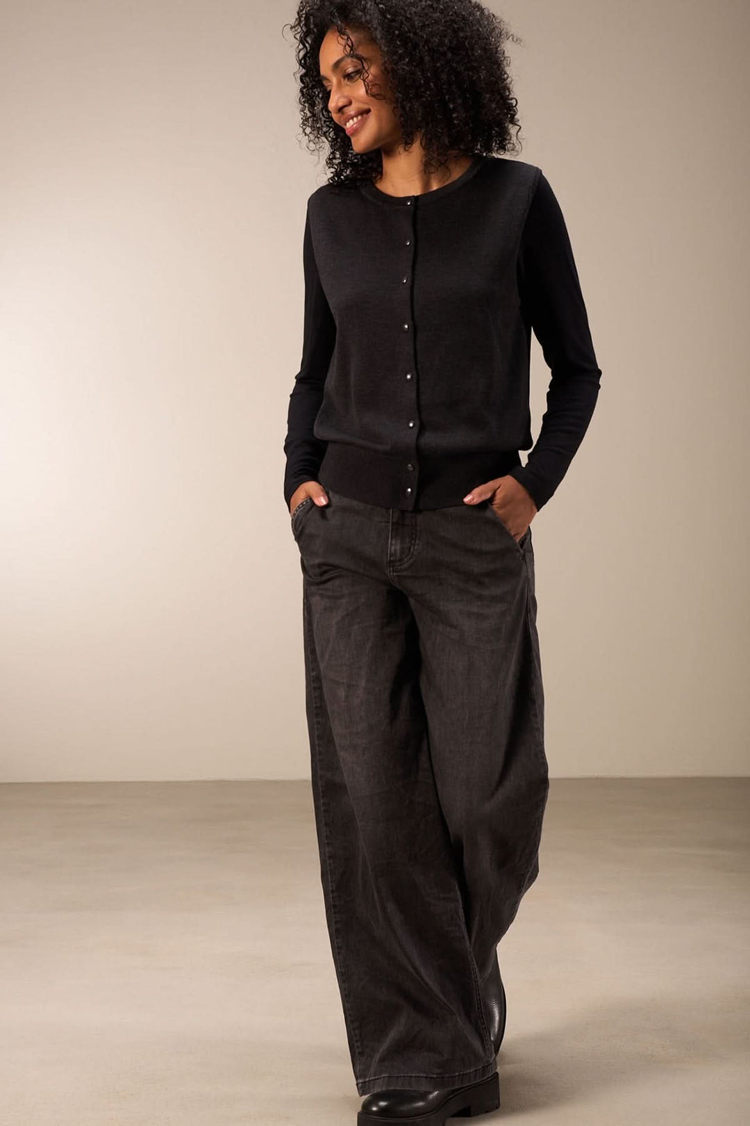Femme souriante portant un cardigan noir boutoné avec un pantalon large, style chic et décontracté.