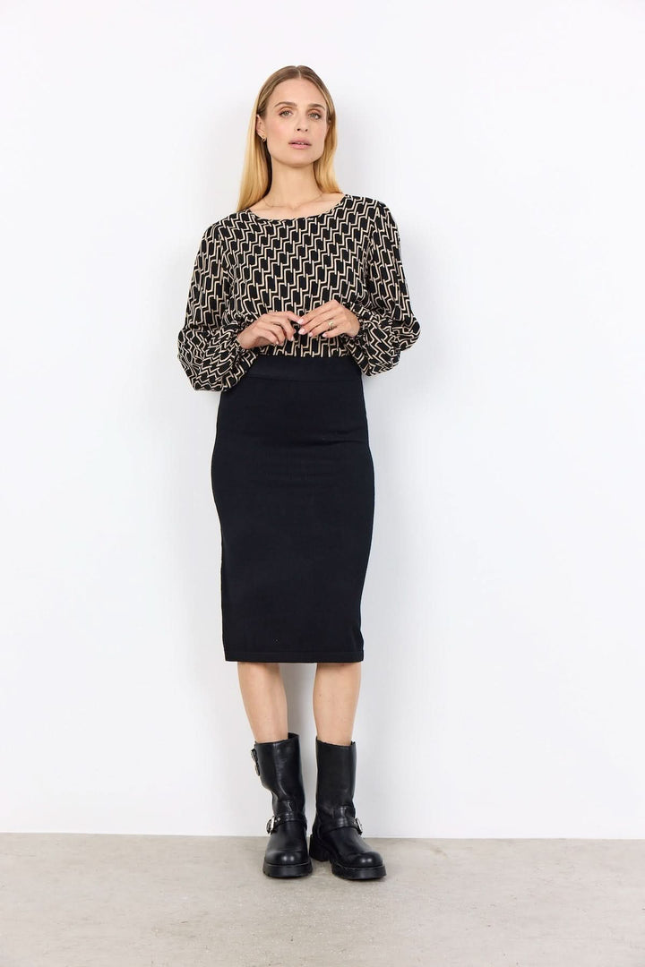 Femme portant une blouse à motif graphique, jupe noire et bottes, look moderne et tendance.