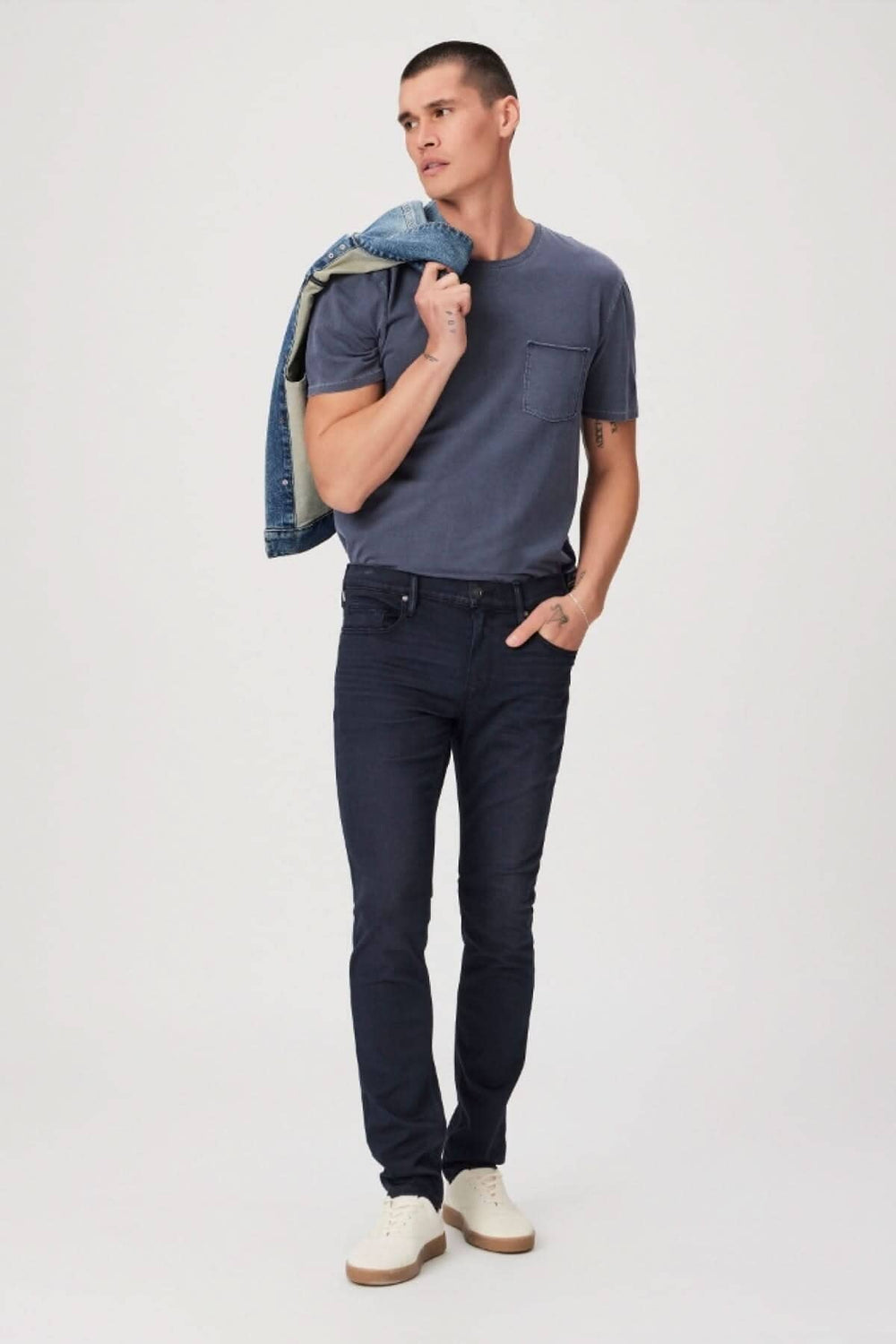 Homme portant un t-shirt gris et un jean foncé, avec une veste en jean sur l'épaule, pose décontractée sur fond neutre.