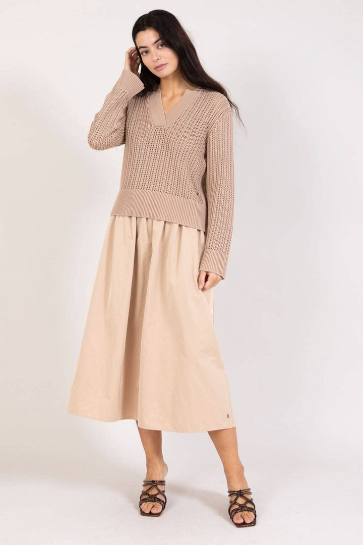 Femme en chandail maille ouverte beige avec encolure en V et jupe beige, pose élégante sur fond neutre.
