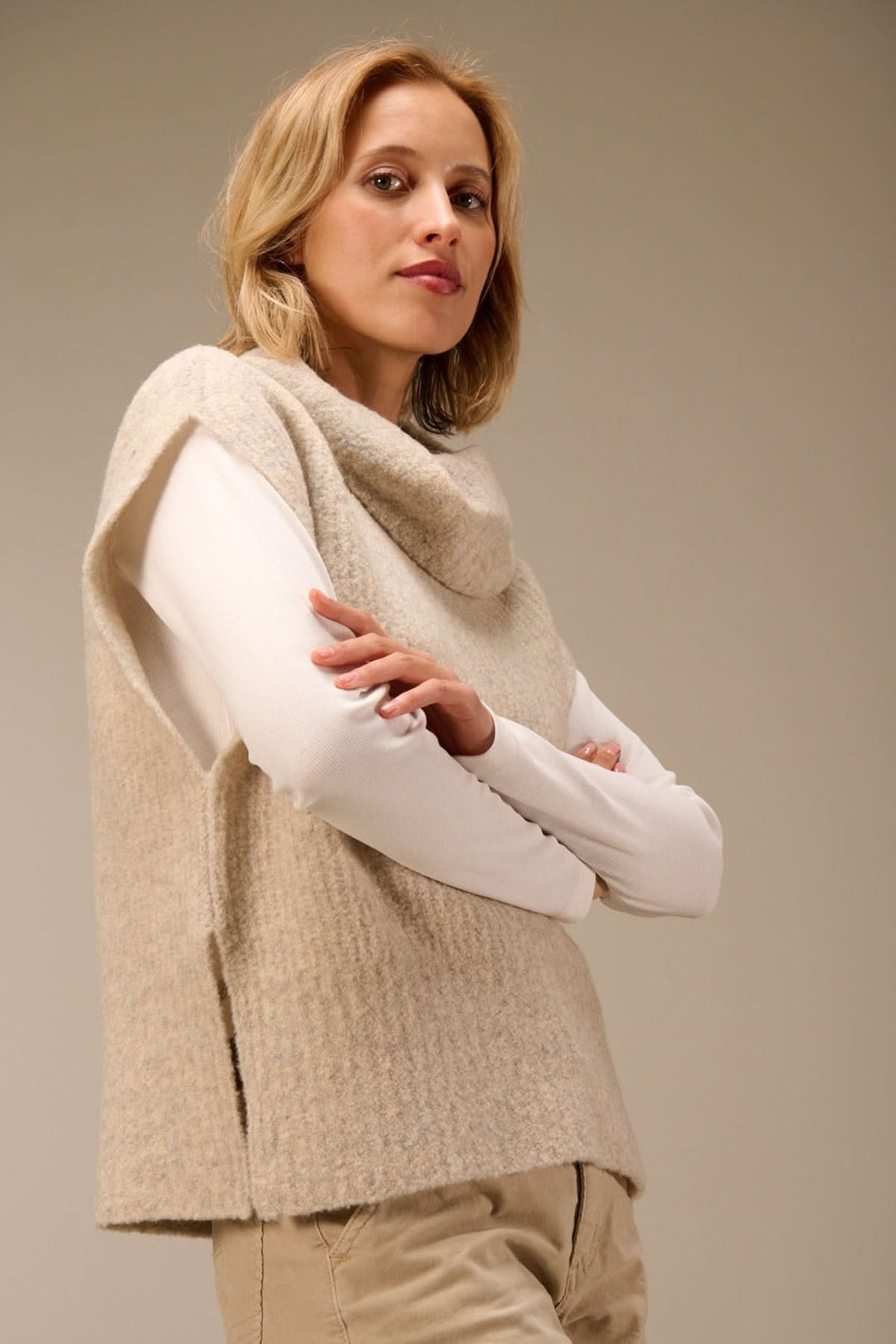 Femme portant un gilet sans manches en maille bouclée à col drapé, superposé sur un t-shirt à manches longues blanc, look élégant.