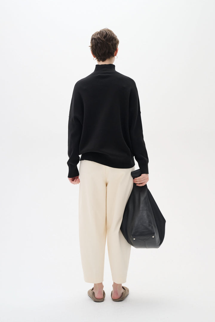 Femme de dos portant un col roulé Tenley noir avec un pantalon blanc, tenant un grand sac noir.
