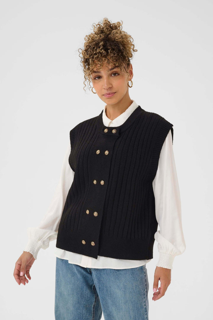 Veste Eroka sans manches en tricot à boutons dorés portée sur chemise blanche en style chic et décontracté, idéale pour mi-saison.