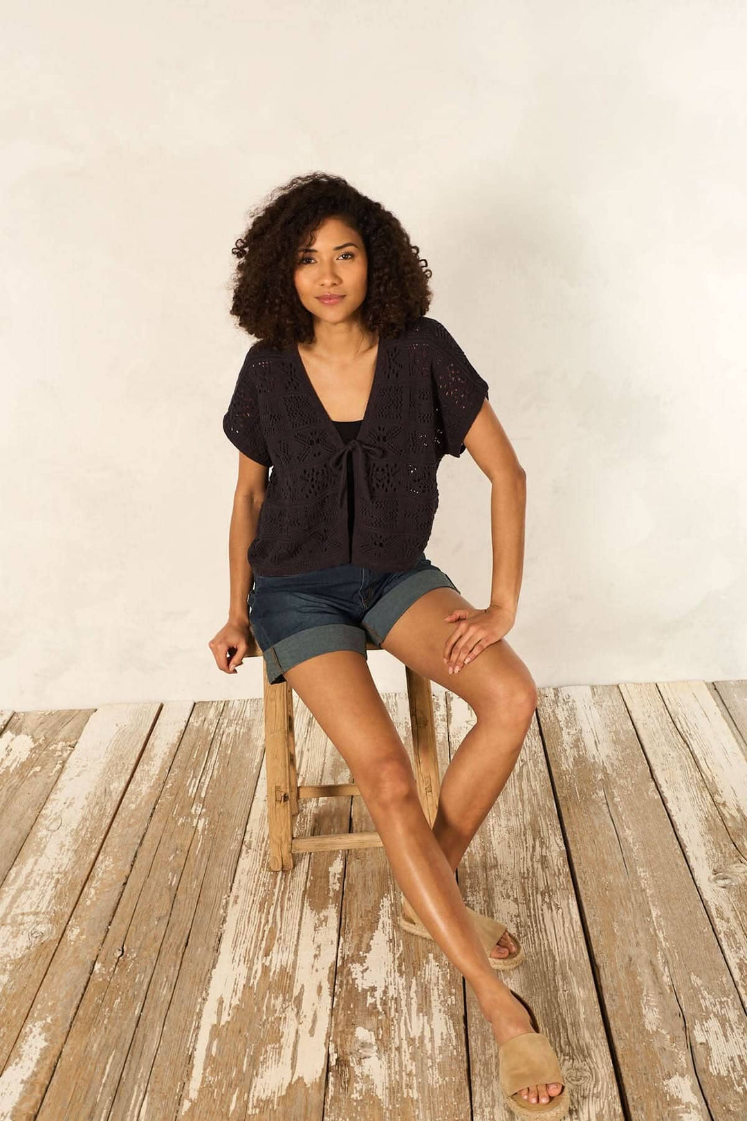 Femme portant un cardigan en maille ajourée à manches courtes avec un ruban, assise sur une chaise en bois.