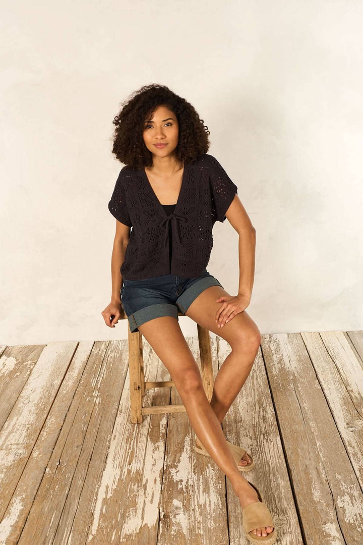 Femme portant un cardigan en maille ajourée à manches courtes avec un ruban, assise sur une chaise en bois.