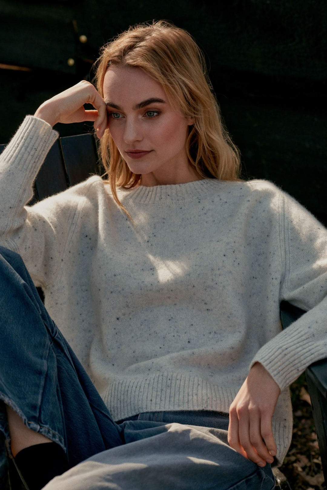 Femme portant un chandail moucheté en cachemire, assise, avec un jean, dans un cadre naturel.