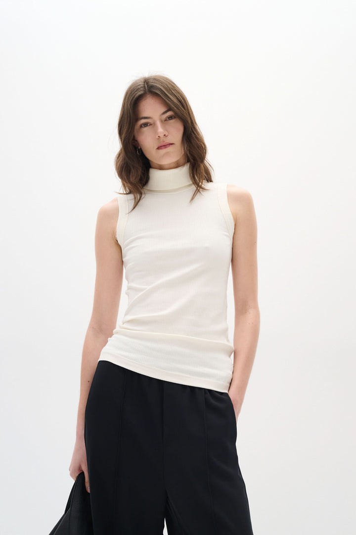Camisole Dagna sans manches à col montant, tissu côtelé, couleur crème, style ajusté, portée avec un pantalon noir.