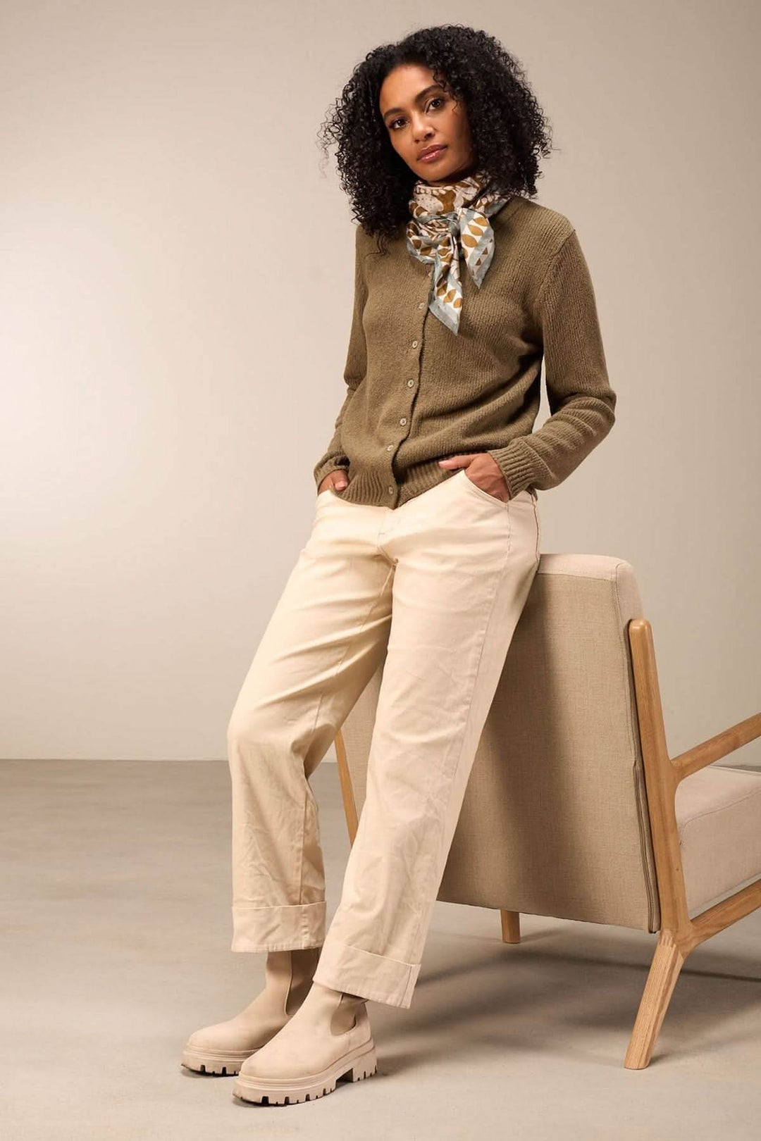 Femme en pull olive avec foulard, pantalon crème et bottes, posant près d'un canapé.