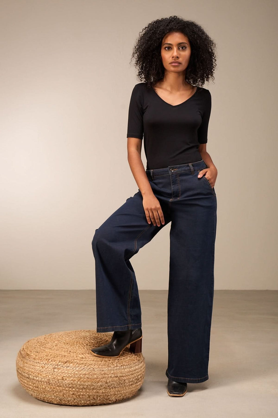 Femme portant un jean coupe large taille mi-haute, associé à un haut noir, sur un pouf en jute.