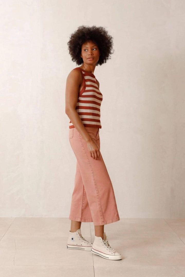 Femme portant un haut à rayures marron et une jupe rose, avec des sneakers blanches, dans un décor neutre.