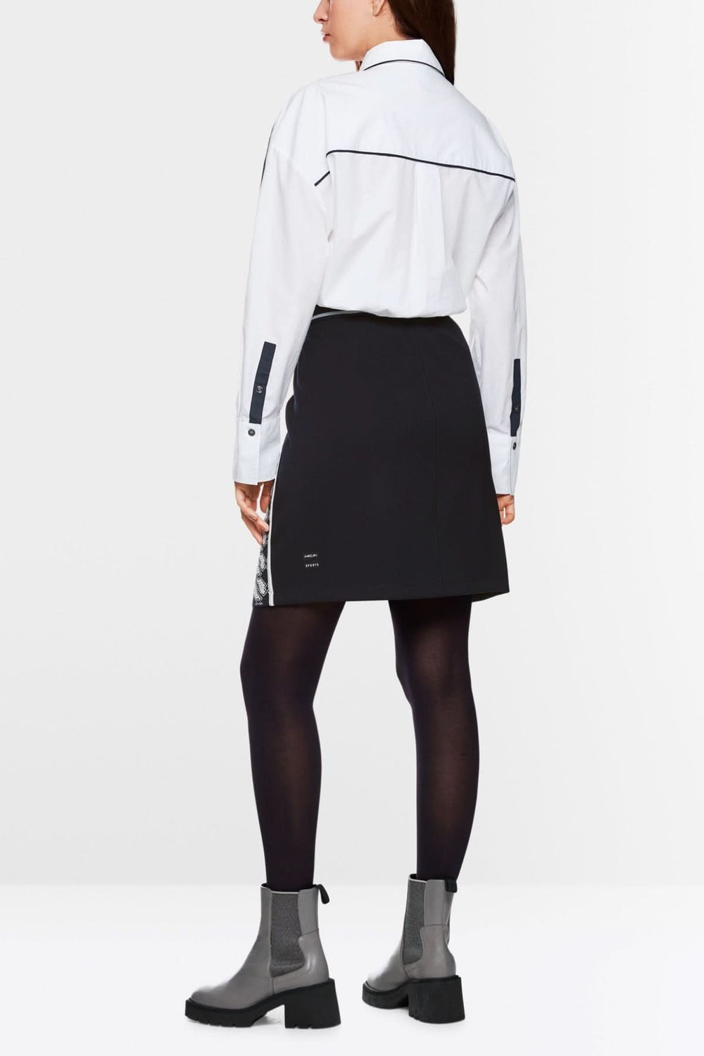 Femme vue de dos portant une jupe en maille noire avec une chemise blanche, combinée avec des bottes élégantes.