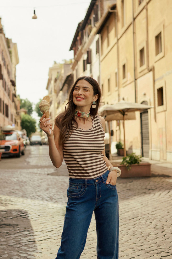 Femme souriante portant une camisole rayée, tenant une glace, dans une rue ensoleillée avec des bâtiments historiques.