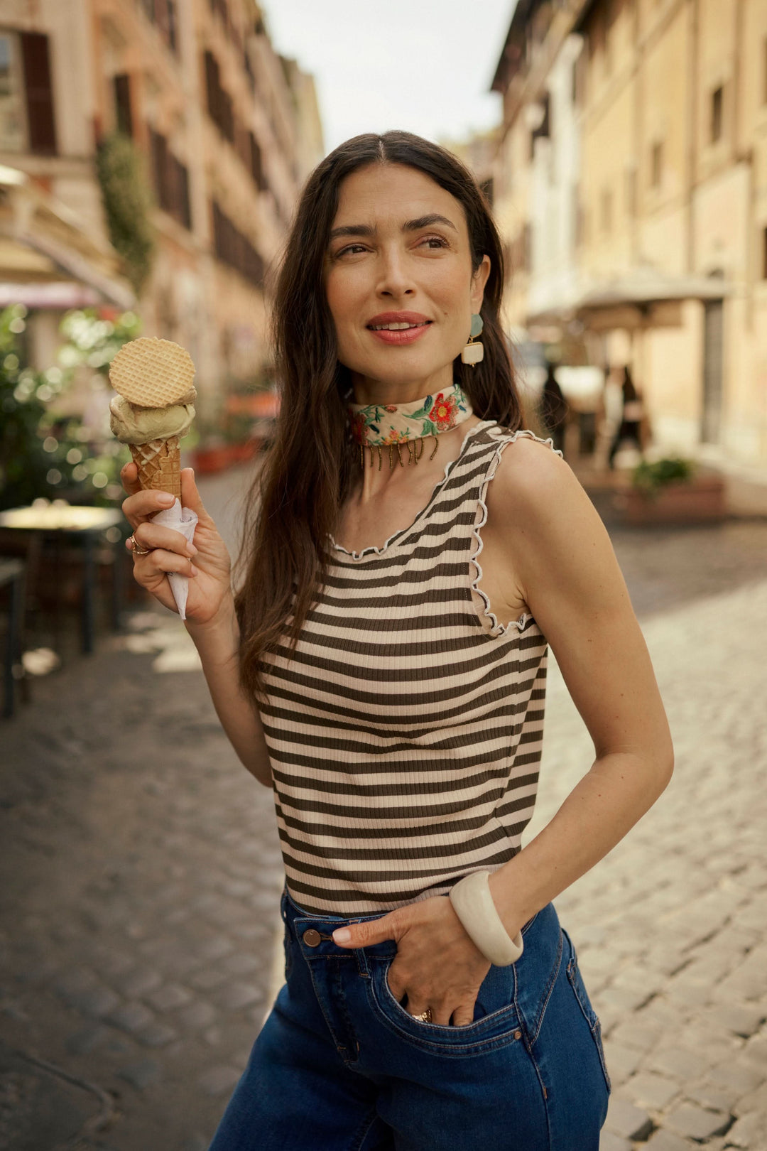 Femme souriante portant une camisole rayée, dégustant une glace dans une rue pittoresque.