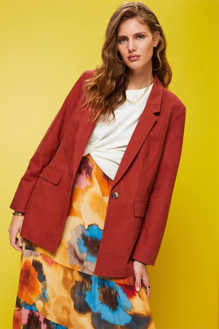 Blazer à simple boutonnage en lin rouge, porté avec une jupe colorée, devant un fond jaune.