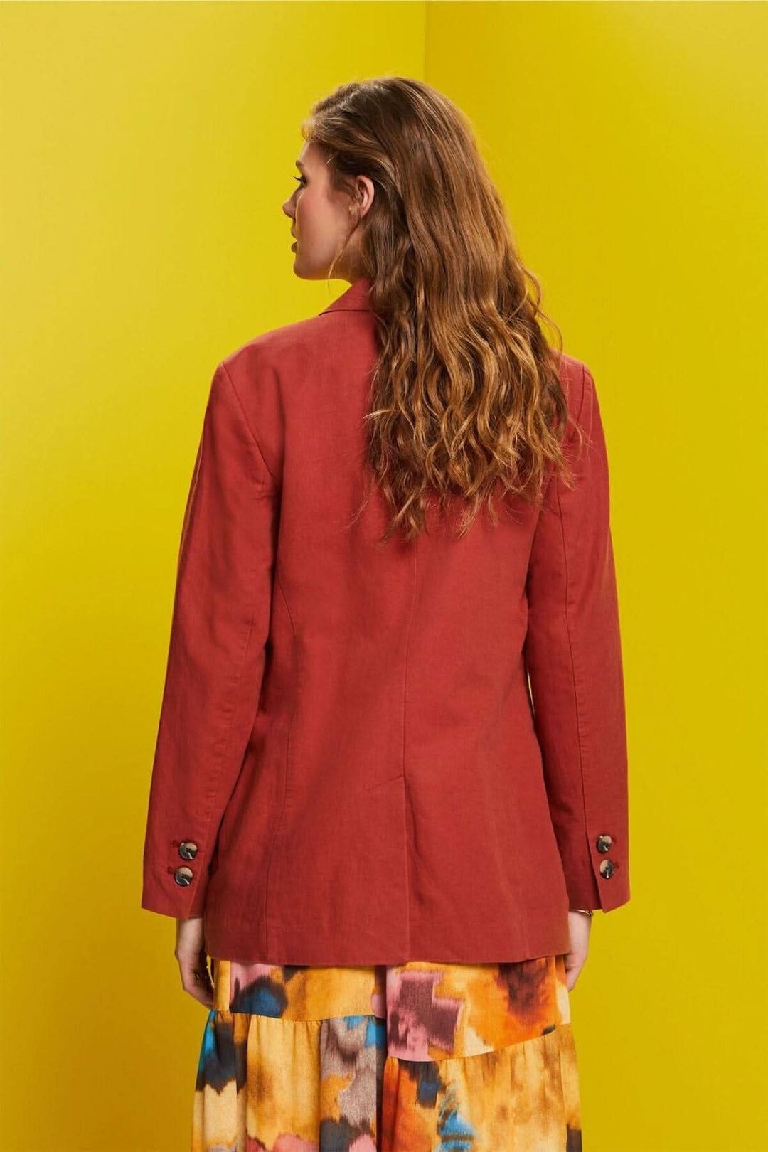 Femme de dos portant un blazer à simple boutonnage rouge, avec col à revers et tissu en lin sur fond jaune.