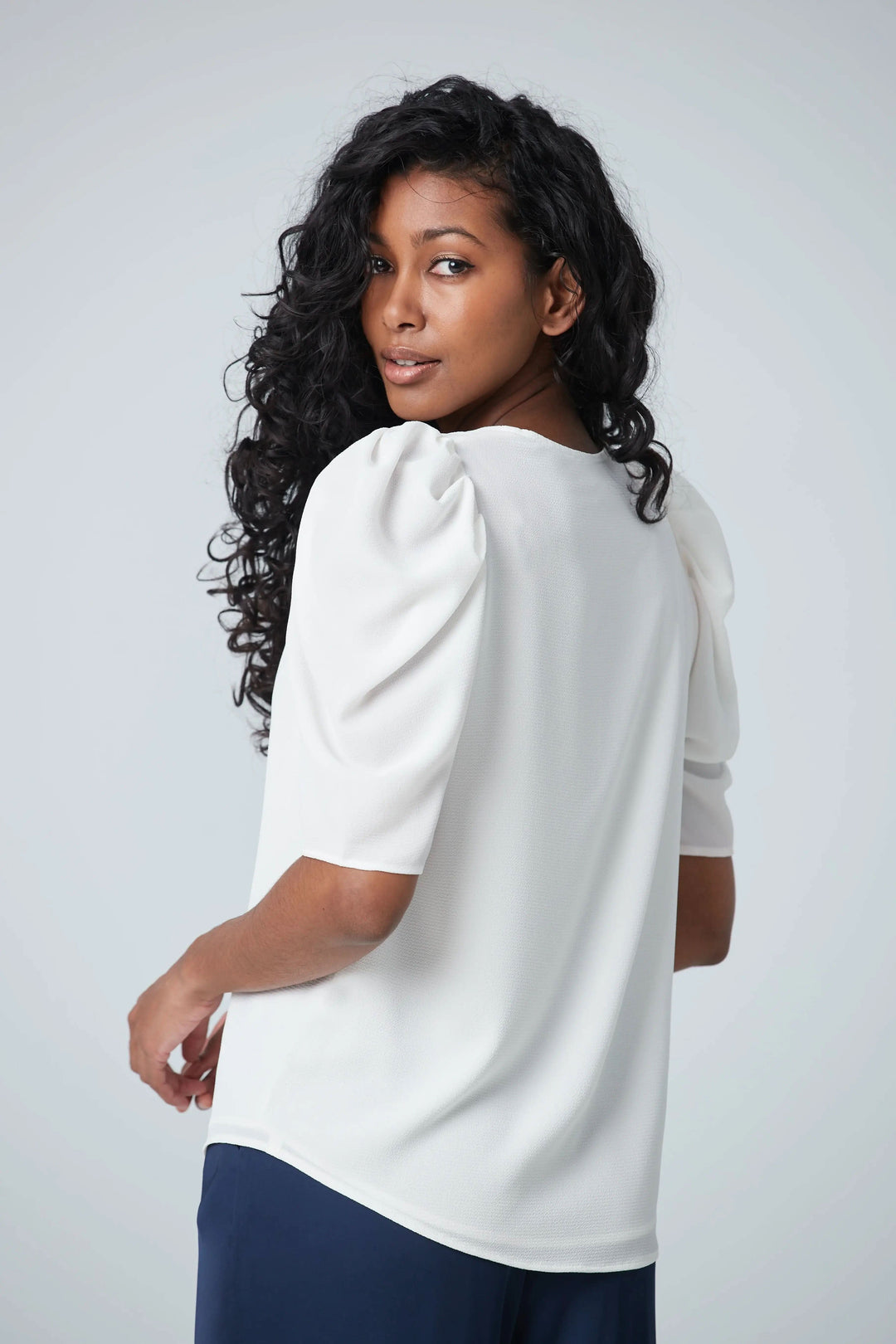 Femme portant une blouse blanche à manches courtes gigots, vue de dos, mettant en valeur un col en V et un tissu léger.