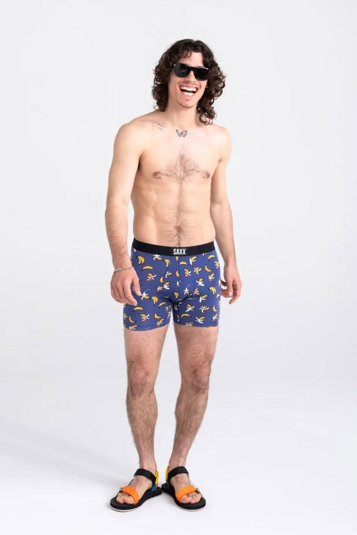 Homme souriant portant des boxers à imprimé de bananes arc-en-ciel, lunettes de soleil et sandales.