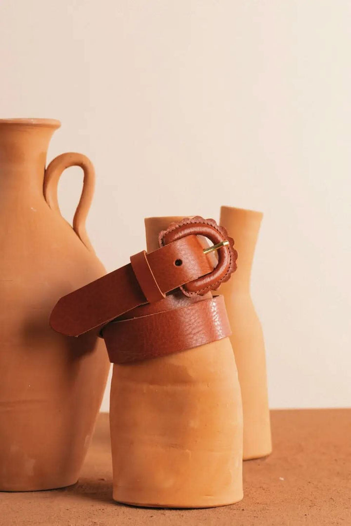 Ceinture Marguerite en cuir acajou avec boucle festonnée, posée sur des vases en terre cuite.