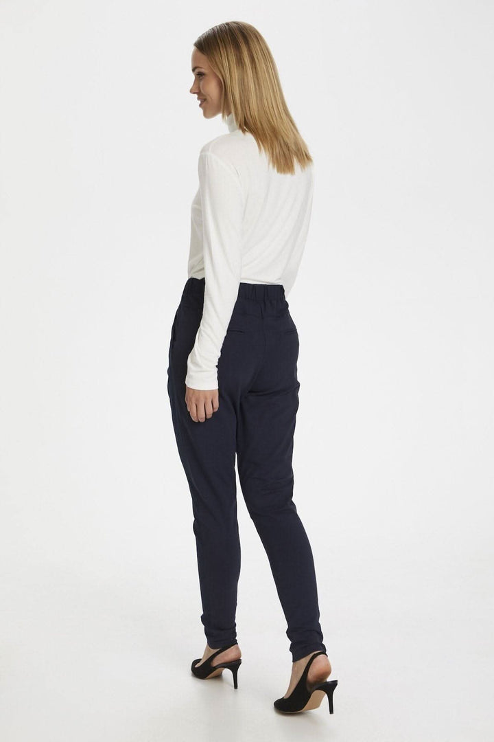 Femme portant un chandail à col roulé blanc et un pantalon noir, vu de dos, sur un fond blanc.