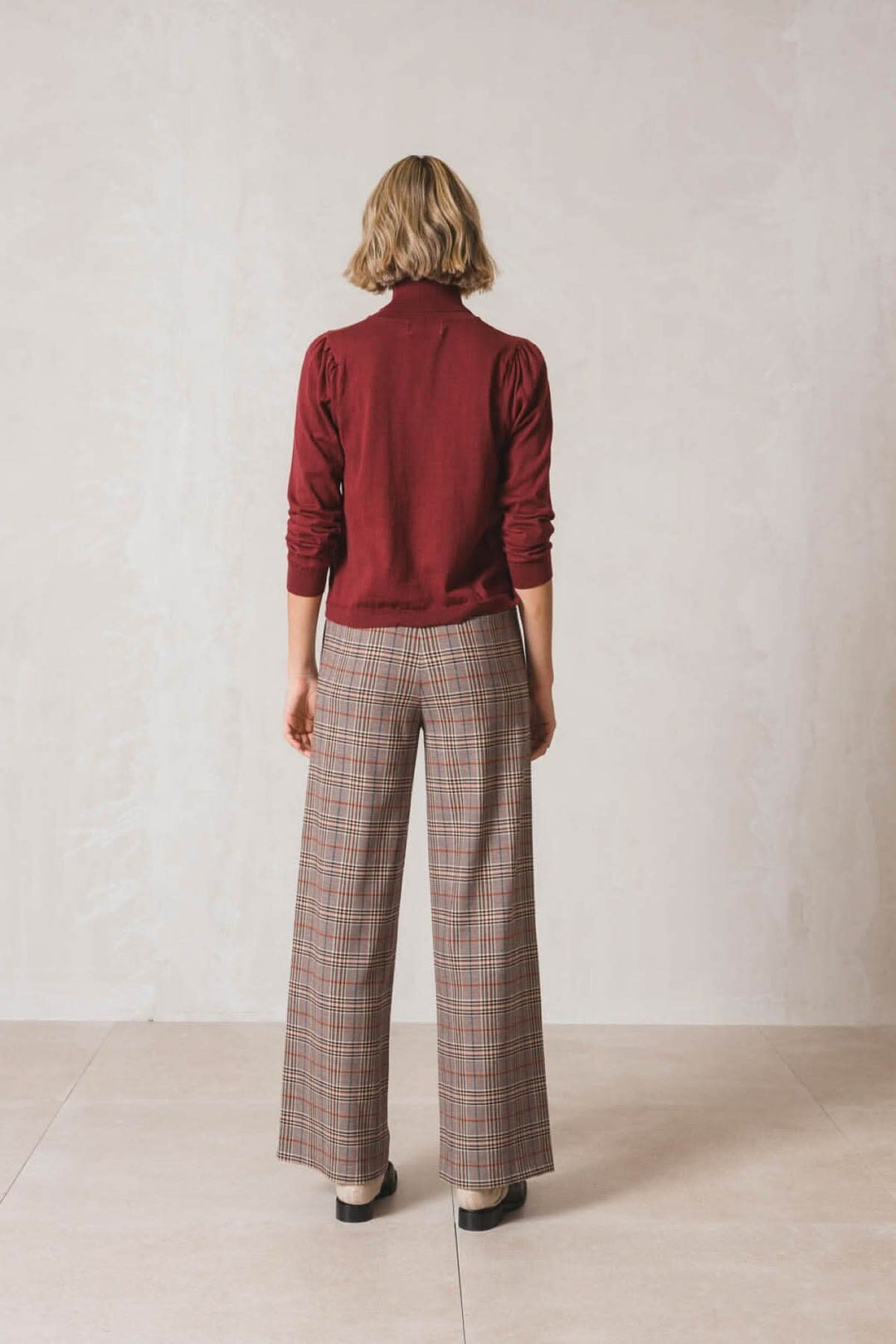 Femme vue de dos portant un chandail col roulé rouge et un pantalon à carreaux, look chic et décontracté.