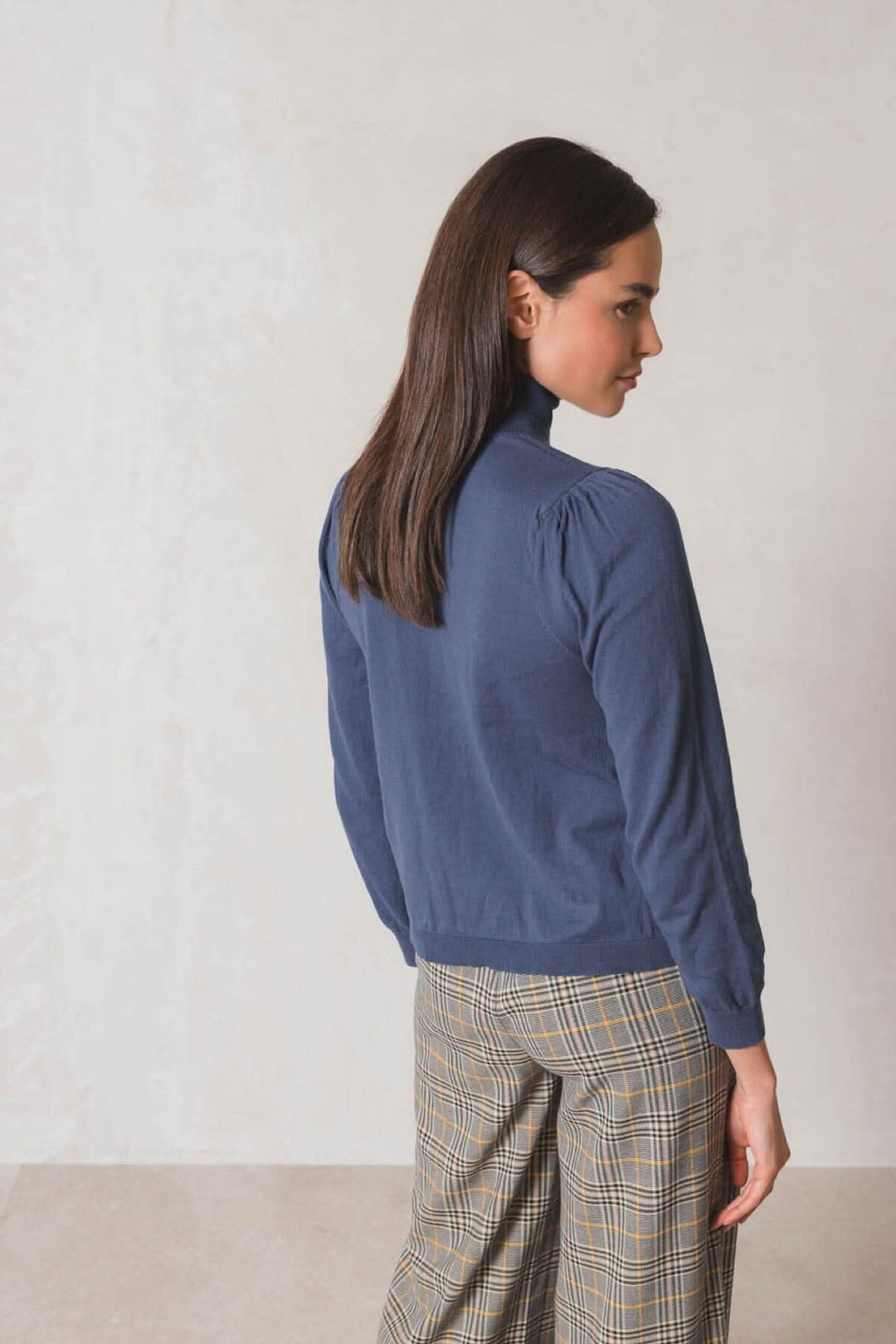 Femme portant un chandail col roulé bleu avec manches longues et plis aux épaules, associé à un pantalon à carreaux.