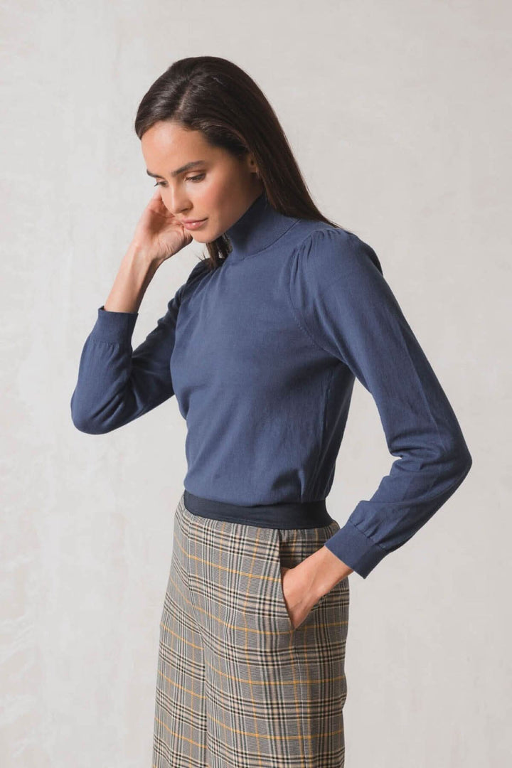 Femme portant un chandail col roulé bleu, manches longues, et jupe à carreaux, style élégant et moderne.