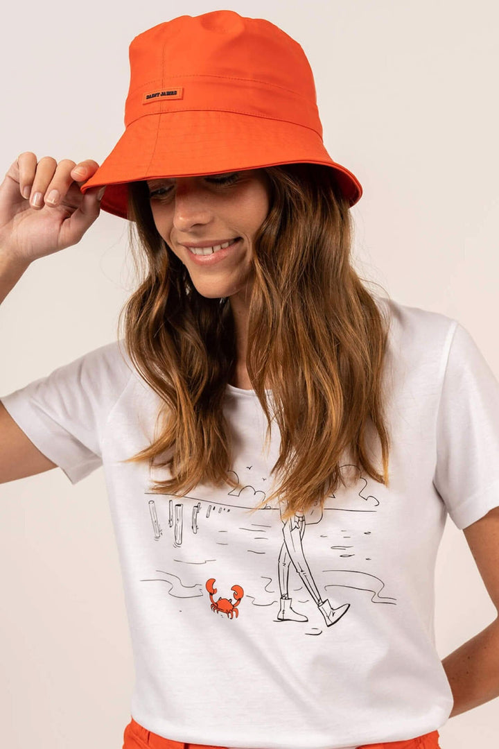 Femme portant un chapeau bob orange sur une plage, avec un t-shirt illustré et des cheveux ondulés.