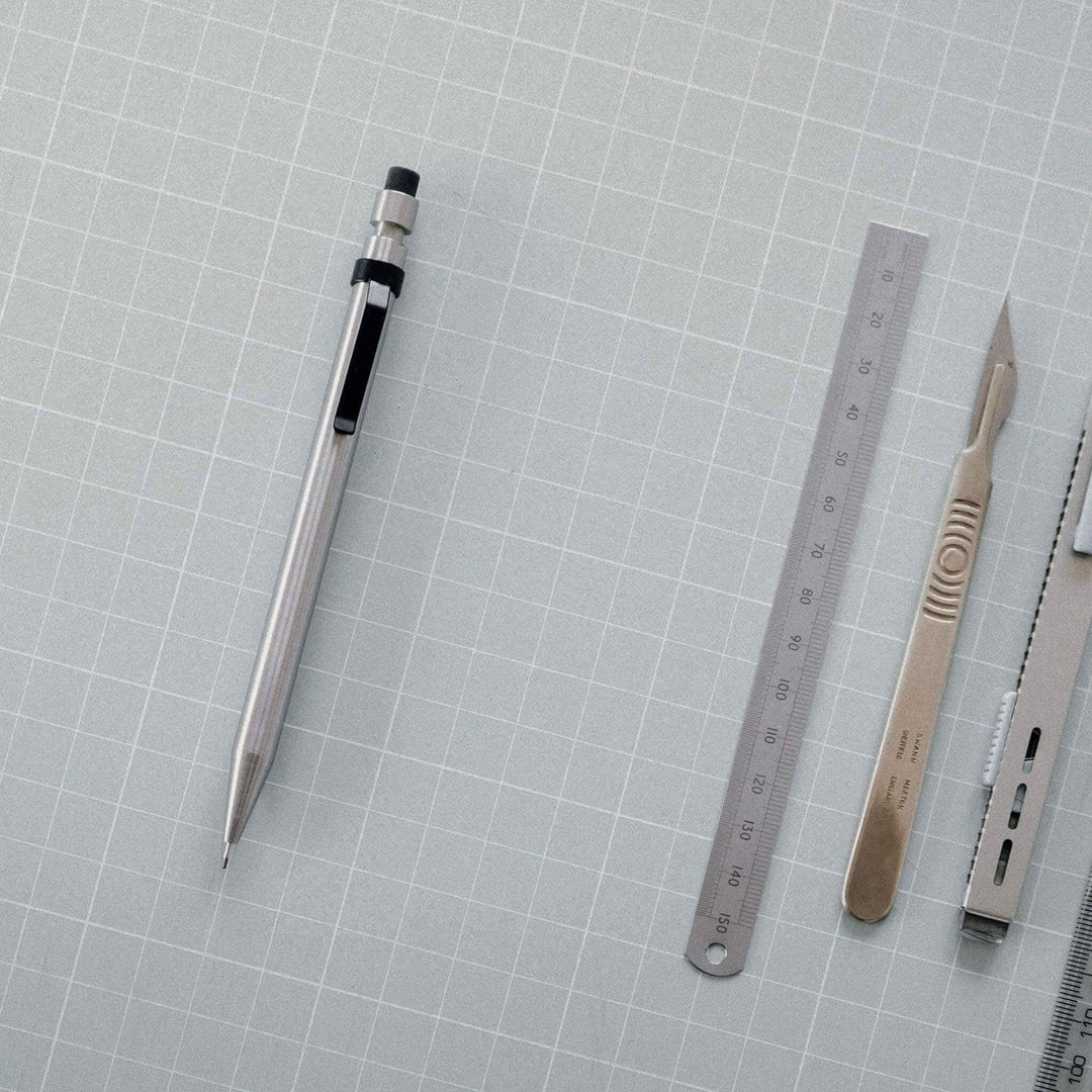 Image de fournitures de bureau incluant un crayon en métal, une règle, un scalpel et d'autres outils sur un fond en quadrillage.