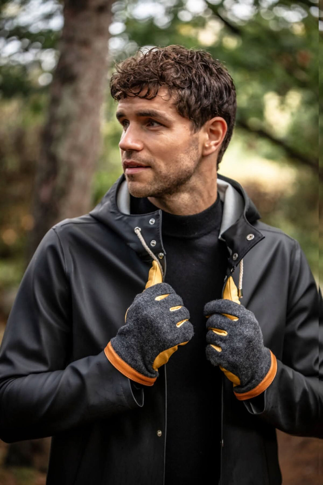 Homme portant des gants en daim et en tricot de laine, sous un manteau noir, dans un cadre naturel.