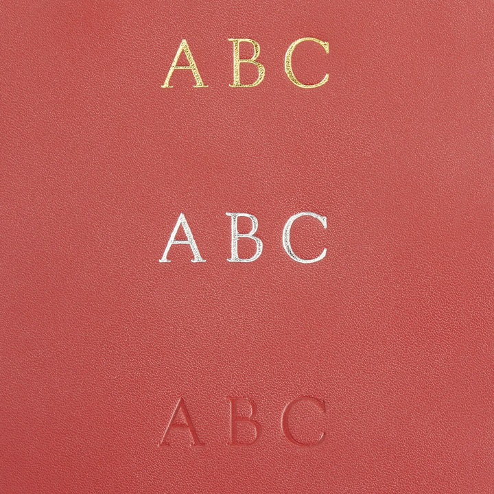 Close-up of three monogram options 'ABC' in gold, silver, and embossed styles on red leather for personalized luggage tags.