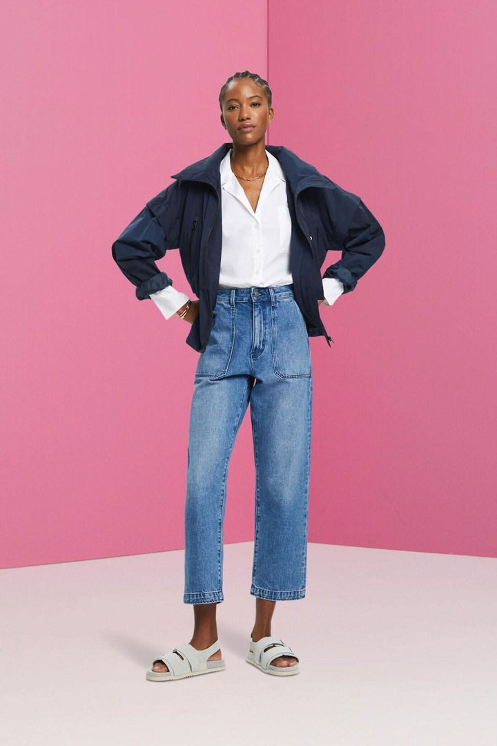 Femme en pantalon Jean coupe Dad, chemise blanche et veste, sur fond rose.