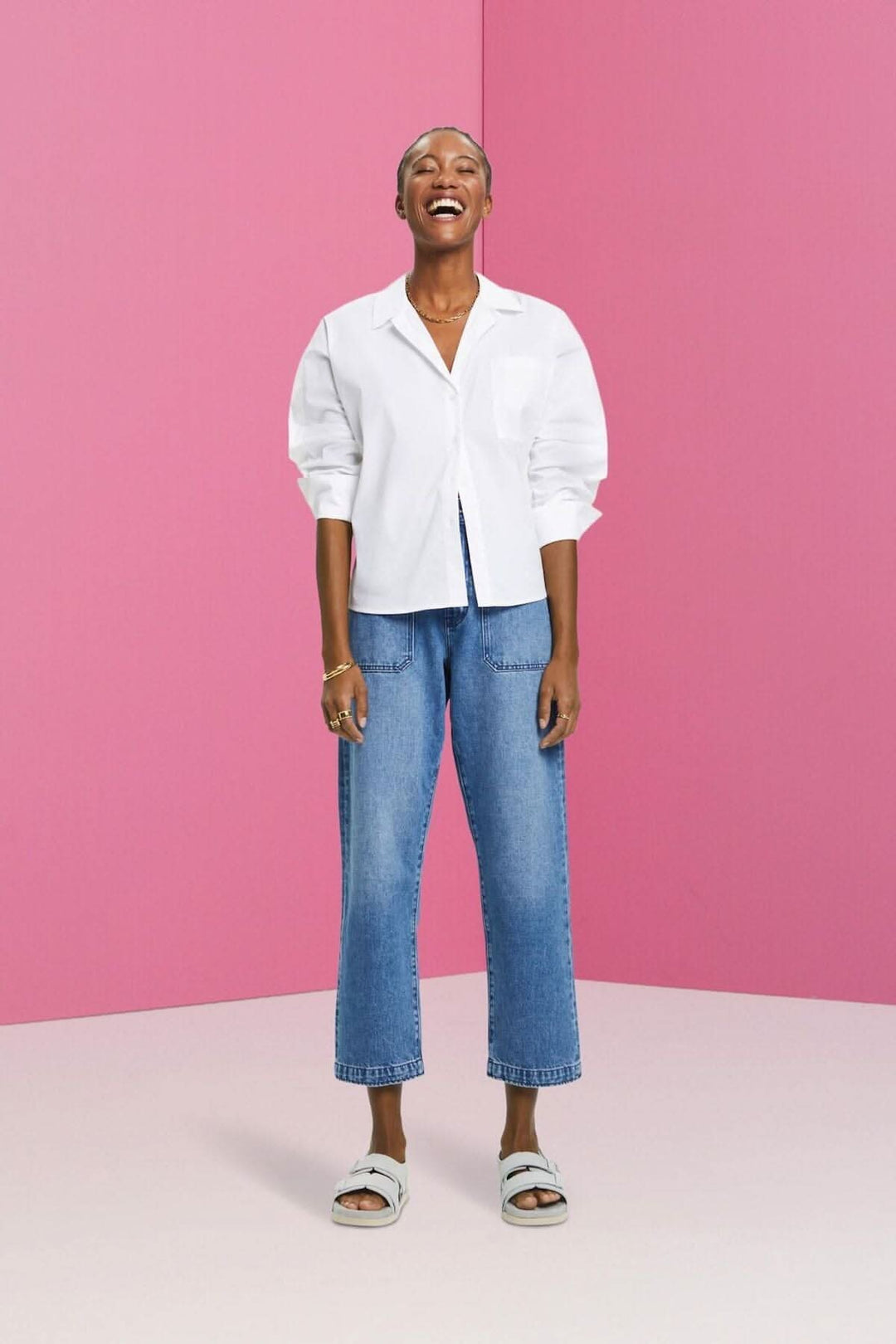 Femme souriante portant une chemise blanche oversize et un jean coupe Dad devant un fond rose.