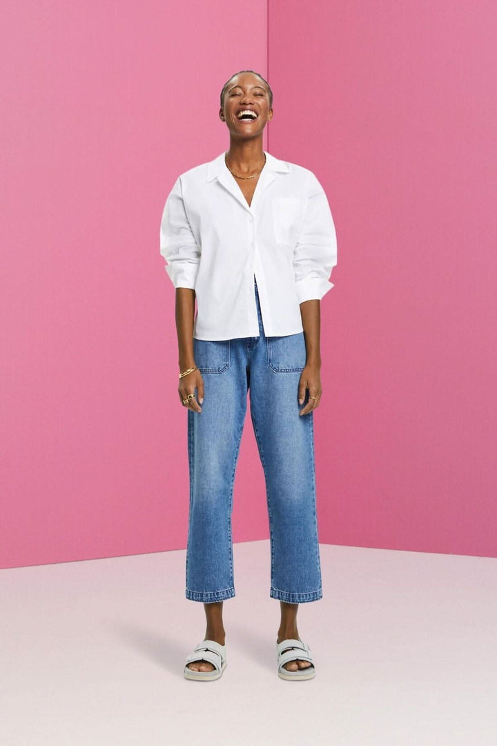 Femme souriante portant une chemise blanche oversize et un jean coupe Dad devant un fond rose.