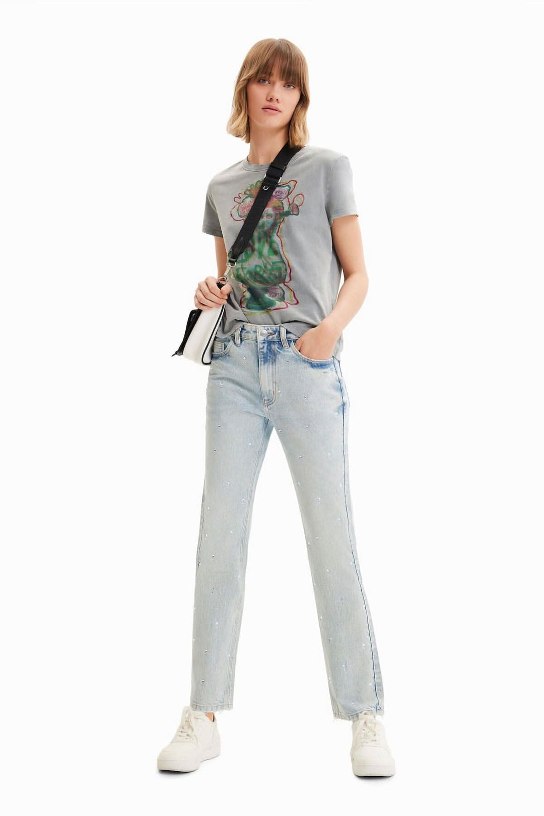 Femme portant un jean droit à strass, tee-shirt gris, et sneakers blanches, pose stylée sur fond blanc.