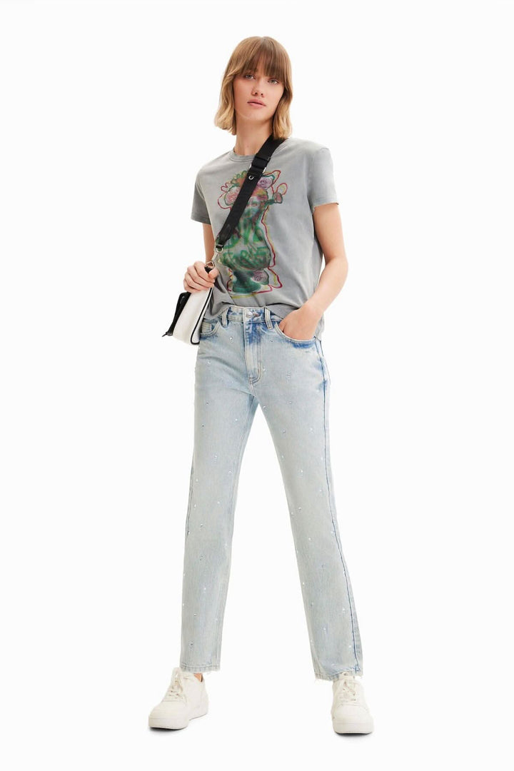 Femme portant un jean droit à strass, tee-shirt gris, et sneakers blanches, pose stylée sur fond blanc.
