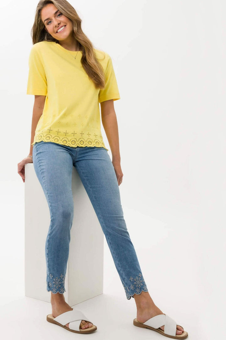 Femme souriante portant un t-shirt jaune et un jean 7/8, avec des sandales blanches, sur fond neutre.