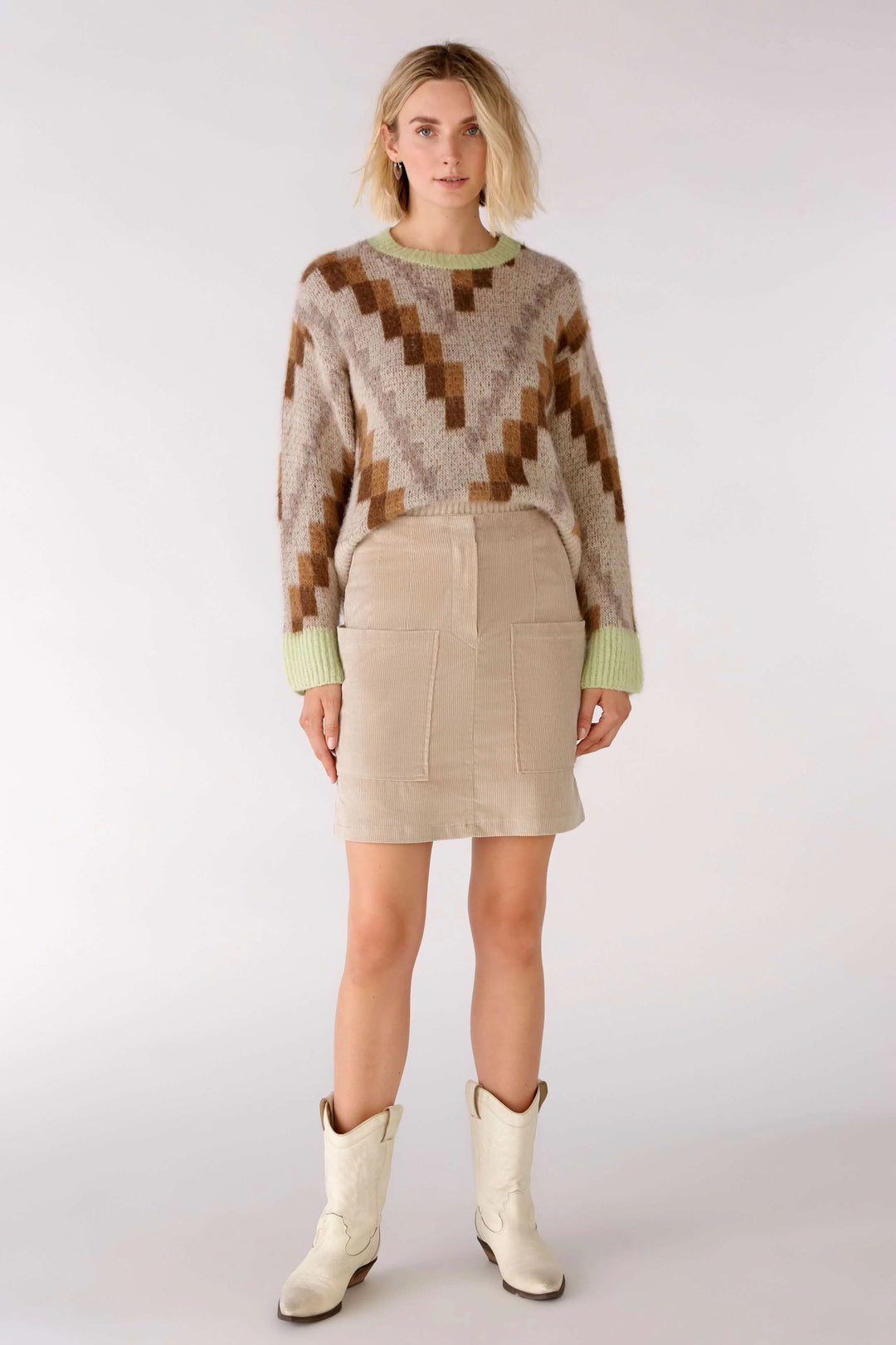 Femme portant une jupe en velours côtelé beige avec des poches, accompagnée d'un pull coloré et de bottes blanches.