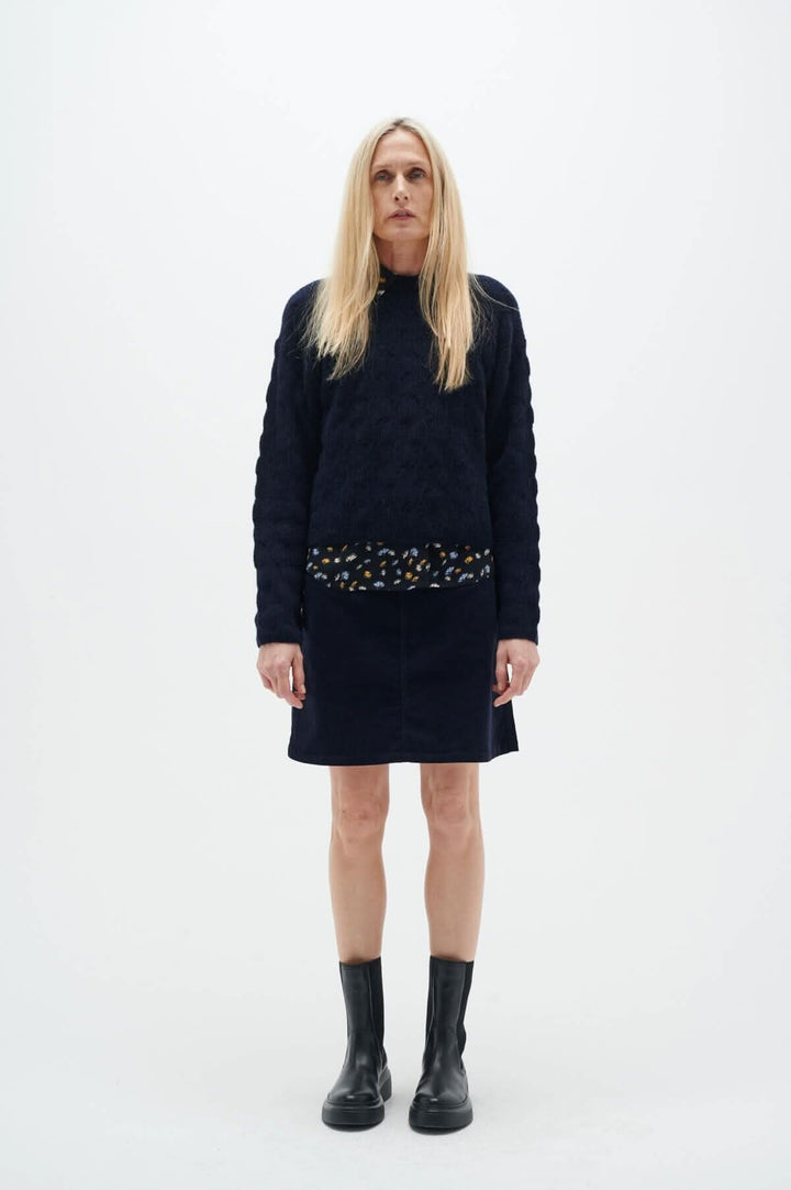 Femme en jupe marine Rylie de Inwear, avec un haut noir, bottes noires, devant un fond blanc.