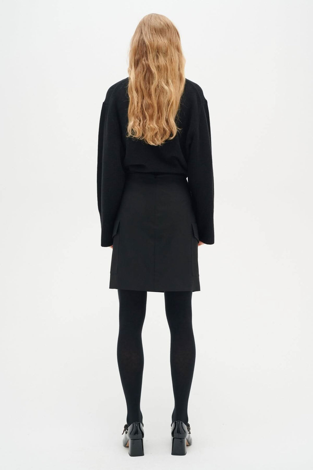 Femme vue de dos, portant une jupe noire avec un chandail ample et des collants noirs.