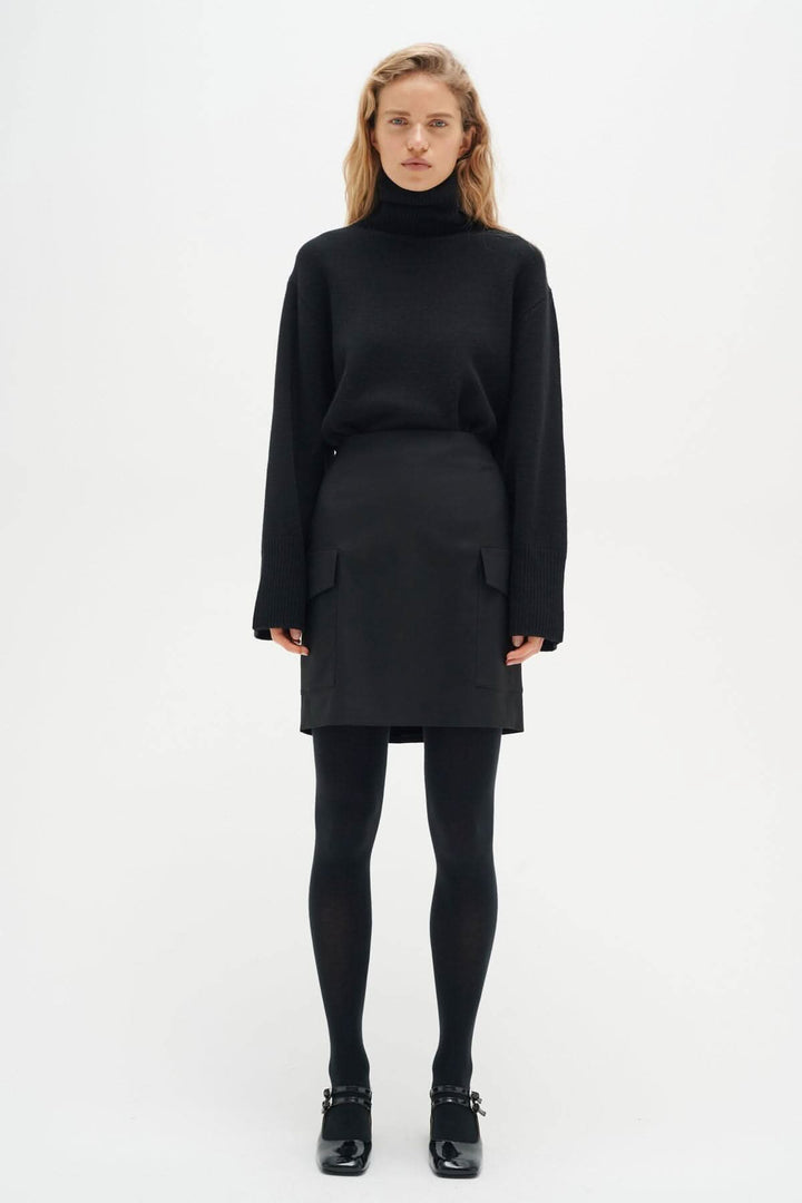 Model wearing a black turtleneck sweater with a matching skirt, styled with black tights and shoes.