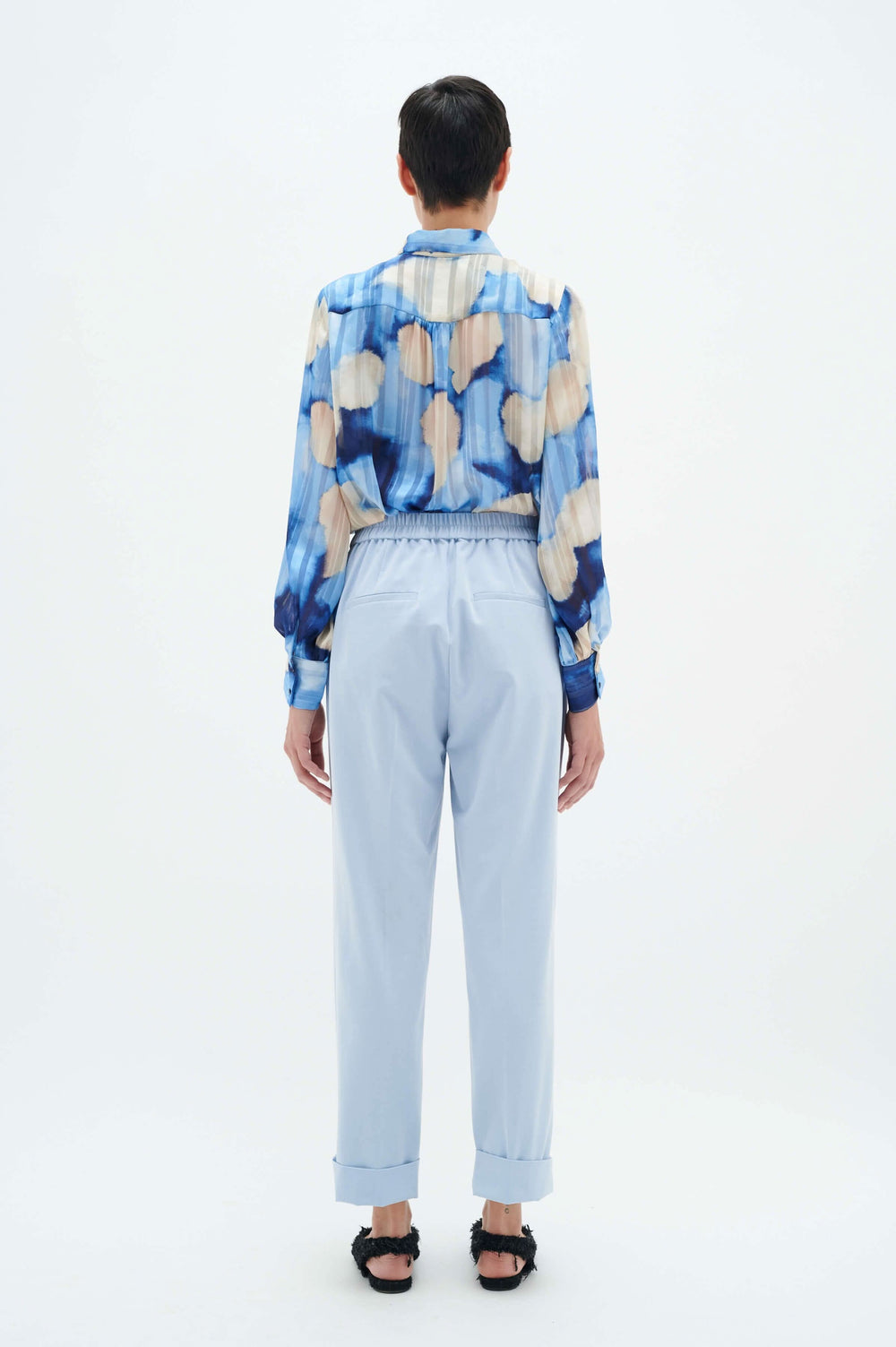 Femme vue de dos portant une chemise bleu à motifs nuage et un pantalon à revers avec des chaussures noires.