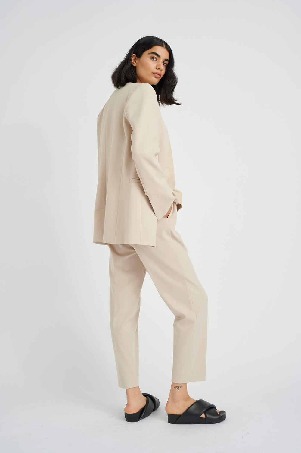 Femme vêtue d'un costume beige moderne avec des pantalons ajustés et des sandales noires, posant de côté.