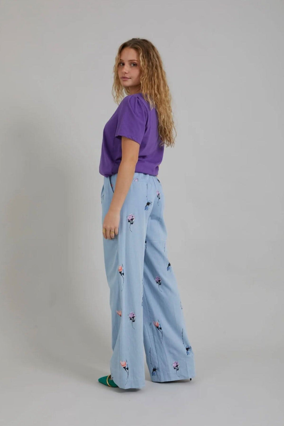 Femme portant un pantalon large en denim floral, haut violet, pose de profil, look décontracté et original.