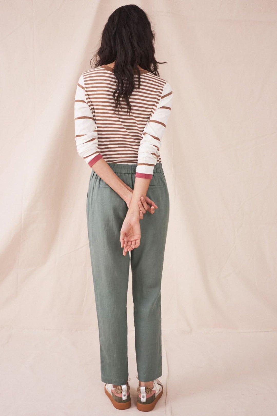 Femme de dos portant le pantalon Maddie en lin vert, associé à un haut rayé, sur fond beige.